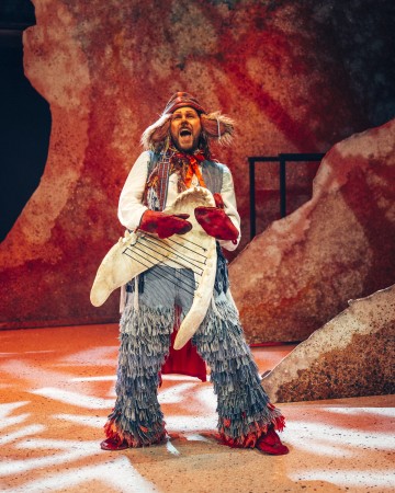 Värikkääseen, höyhenpehmeään pukuun pukeutunut esiintyjä seisoo lavalla ja pitää kädessään hainmuotoista rekvisiittaa, jossa on nuottiviivoja muistuttavia viivoja. Taustalla on abstrakteja, kuvioituja maisemia lämpimillä sävyillä.