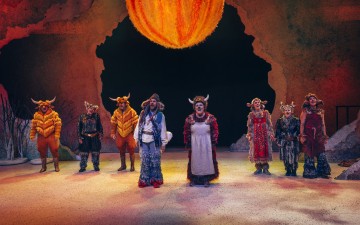 Kahdeksan esiintyjää värikkäissä, omituisissa puvuissa ja sarvissa seisoo lavalla suuren, oranssina hehkuvan pallon alla, luolamaisen taustan edessä, ikään kuin keskellä teatteriesitystä.