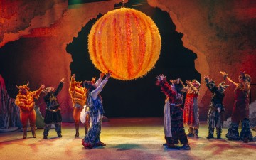 Värikkäisiin, karvaisiin pukuihin ja sarviin pukeutuneet esiintyjät nostavat kätensä kohti suurta, oranssina hehkuvaa palloa, joka on ripustettu näyttämön yläpuolelle, jossa on kivinen, kuvioitu tausta. Kohtaus on eläväinen ja teatraalinen.