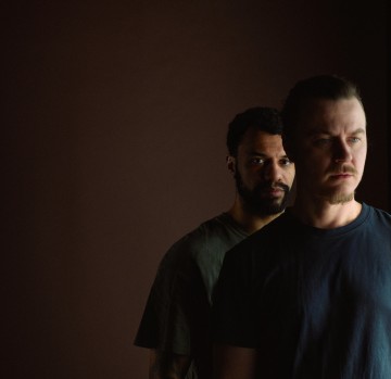 Kaksi miestä seisoo lähekkäin tummaa taustaa vasten. Toinen mies on hieman toisen takana ja vasemmalla puolella, molemmilla on neutraali ilme ja yllään tummat t-paidat.
