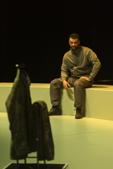 Harmaapukuinen mies istuu matalalla, vaalealla näyttämötasanteella ja katsoo kohti naamiotakkia, joka on esillä etualalla dramaattisessa valaistuksessa.