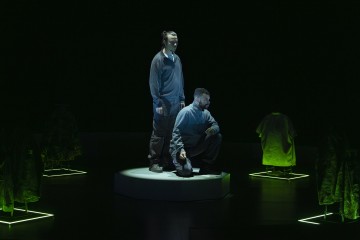 Kaksi ihmistä seisoo pienellä pyöreällä korokkeella dramaattisessa valaistuksessa, ja heitä ympäröivät pimeällä näyttämöllä vihreällä valolla valaistuihin telineisiin ripustetut vaatteet.