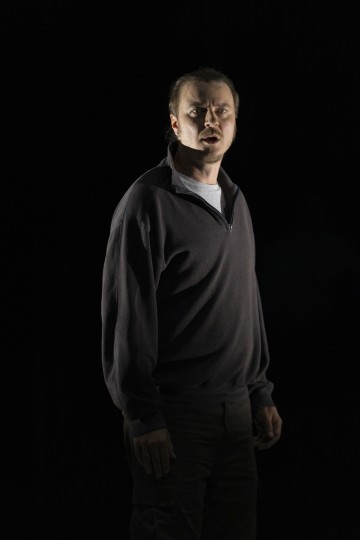 Mies seisoo pimeässä tilassa, jota valonheitin valaisee. Hänellä on tumma villapaita ja housut, kasvoilla vakava tai intensiivinen ilme, katse hieman sivulle.