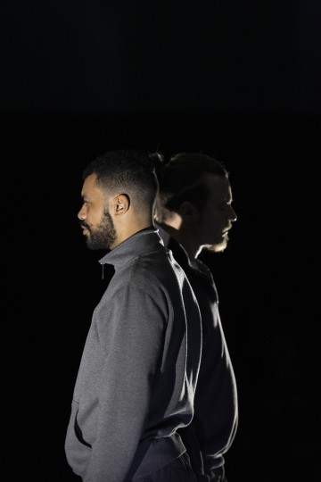 Kaksi miestä seisoo selkä vastakkain pimeässä ympäristössä, molemmat vastakkaisiin suuntiin. Toinen on etualalla, sivulta valaistuna, kun taas toinen on taustalla, osittain varjossa. Molemmilla on tummat vaatteet.