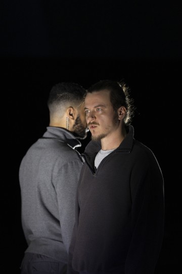 Kaksi miestä seisoo lähekkäin; toinen katsoo eteenpäin vakavalla ilmeellä, kun taas toinen katsoo poispäin, ja vain hänen päänsä sivu ja takaraivo ovat näkyvissä. Molemmilla on yllään tummat villapaidat mustaa taustaa vasten.