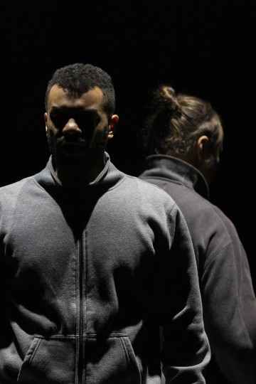 Kaksi vetoketjulliseen takkiin pukeutunutta ihmistä seisoo selkä vastakkain hämärässä valaistuksessa; toinen katsoo eteenpäin varjossa, kun taas toinen kääntyy poispäin, kasvot osittain piilossa. Tausta on täysin pimeä.