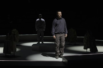 Kaksi ihmistä seisoo valonheittimien alla hämärästi valaistulla näyttämöllä, kumpikin erillisillä korokkeilla, ja heitä ympäröivät telineissä olevat sotilastyyliset takit; tunnelma on jännittynyt ja dramaattinen.