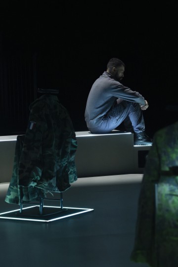 Harmaisiin vaatteisiin pukeutunut mies istuu yksin valkoisella alustalla hämärässä huoneessa. Etualalla, alhaalta valaistulla jalustalla on sotilasmaastotakki, jossa on Ranskan lippulappu.