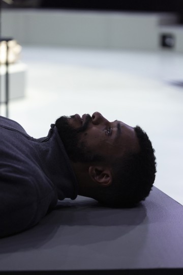 Mies, jolla on parta ja lyhyet hiukset, makaa selällään tasaisella alustalla ja katsoo ylöspäin. Hänellä on yllään tumma vetoketjullinen takki, ja tausta on pehmeästi sumentunut neutraaleilla sävyillä.