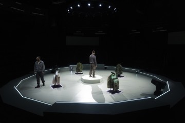 Kaksi ihmistä seisoo pyöreällä, futuristisella näyttämöllä, jossa toinen on keskikorokkeella. Neljä eri asuihin pukeutunutta mallinukkea on sijoitettu ympäri näyttämöä, joka on hämärästi valaistu valonheittimillä.