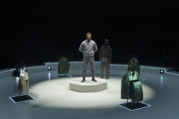 Mies seisoo keskikorokkeella hämärästi valaistulla pyöreällä näyttämöllä. Toinen henkilö seisoo selkä menosuuntaan. Heidän ympärillään on naamiotakkeja esillä telineissä, ja taustalla olevat istuimet ovat tyhjiä.