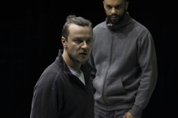 Kaksi harmaisiin vetoketjutakkeihin pukeutunutta miestä seisoo lähekkäin; edessä olevalla miehellä on vakava ilme ja suu hieman auki, kun taas takana oleva mies katselee osittain varjossa tummalla taustalla.