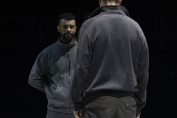 Harmaaseen fleecetakkiin pukeutunut mies seisoo toista henkilöä vastapäätä kädet selän takana. Kohtaus on hämärästi valaistu, ja tausta on tumma, joten kaksi hahmoa ovat pääkohteena.