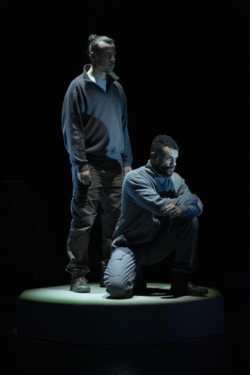 Kaksi miestä, joista toinen seisoo ja toinen polvistuu, ovat valonheittimen valaisemina pimeällä näyttämöllä. Molemmilla on yllään rennot, neutraalin väriset vaatteet, ja he näyttävät vakavilta ja mietteliäiltä.