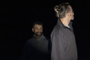 Kaksi tummiin vaatteisiin pukeutunutta miestä seisoo hämärässä ympäristössä; toinen katsoo neutraalilla ilmeellä eteenpäin, kun taas toinen kääntyy poispäin ja katsoo sivulle. Molempia korostaa pehmeä, keskittynyt valo.