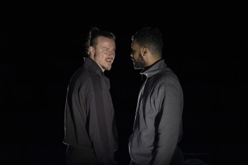 Kaksi miestä seisoo lähellä toisiaan vastakkain pimeässä ympäristössä, molemmilla on tummat takit yllään. Toinen mies katsoo toista intensiivisellä ilmeellä, kun taas toinen katsoo häntä neutraalilla ilmeellä.