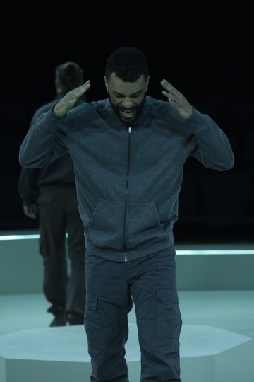 Mies, jolla on harmaa vetoketjullinen huppari ja rahtitaskuhousut, seisoo valaistulla näyttämöllä, nostaa kätensä päänsä eteen ja näyttää huutavan, kun taas toinen henkilö seisoo taustalla kasvot poispäin.