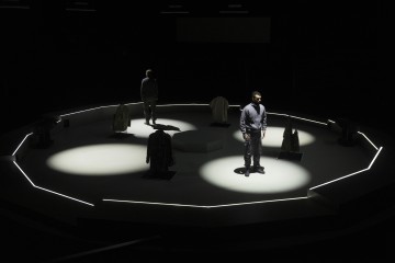 Kaksi ihmistä seisoo hämärästi valaistulla pyöreällä näyttämöllä, jota ympäröivät valonheittimet, ja heidän ympärillään on useita vaatekappaleita mallinukeilla tai jalustoilla. Tausta on tumma, mikä korostaa valaistua aluetta.