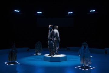 Kaksi ihmistä seisoo selkä toisensa vasten pyöreällä alustalla, jota ympäröivät valaistut vaatteet pimeässä huoneessa, jossa on sininen valaistus. Neljä tyhjää asua on esillä telineissä heidän ympärillään.