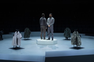 Kaksi ihmistä seisoo valkoisella alustalla eri suuntiin hämärässä huoneessa, jota ympäröi neljä nukkea, joilla on erilaisia sotilastyylisiä maastotakkeja. Taustalla näkyy yleisön istumapaikkoja.