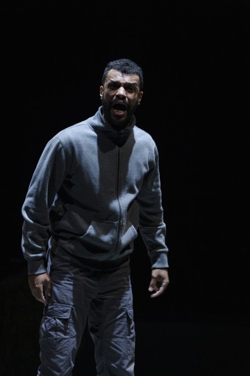 Harmaaseen vetoketjulliseen takkiin ja lasti-housuihin pukeutunut mies seisoo valonheittimen alla ja näyttää huutavan tai puhuvan kiihkeästi, vakavalla ilmeellä, tummaa taustaa vasten.