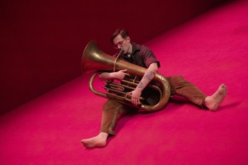 Paljasjalkainen henkilö, jolla on silmälasit ja tatuoinnit, istuu kirkkaan vaaleanpunaisella lattialla ja soittaa suurta messinkitubaa. Hänen jalkansa ovat ojennetut, ja hän näyttää keskittyneen soittimeen.