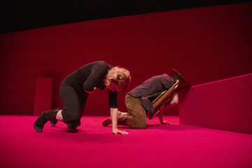 Kaksi ihmistä polvistuu kirkkaanpunaisella näyttämöllä; toinen pitää vatsaansa ja nojaa eteenpäin, kun taas toinen painaa päätään ja vaskisoitinta harkkoa vasten. Molemmat näyttävät näyttelevän dramaattisessa esityksessä.