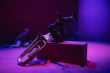 Henkilö makaa selällään palikan päällä, jalat koholla, tuuban vieressä dramaattisessa vaaleanpunaisessa ja sinisessä valaistuksessa. Toinen henkilö polvistuu lattialla taustalla minimalistista lavastusta vasten.