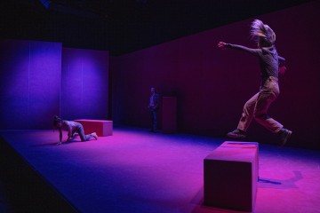 Kolme ihmistä esiintyy hämärästi valaistulla näyttämöllä, jossa on violetti ja vaaleanpunainen valaistus; yksi henkilö hyppää ilmassa palikoiden välissä, toinen polvistuu lattialla, ja kolmas seisoo taustalla lähellä seinää.