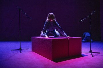 Henkilö polvistuu punaisella korokkeella kahden mikrofonin välissä, dramaattisessa violetissa ja vaaleanpunaisessa valaistuksessa, ja toinen henkilö näkyy osittain lattialla taustalla.