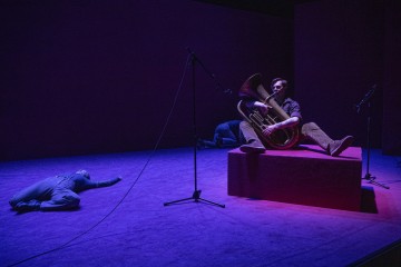 Hämärästi valaistulla lavalla, jossa on violetti valaistus, näkyy lattialla makaava henkilö ja lavalla istuva henkilö, joka soittaa tuubaa, ja heidän ympärillään on useita mikrofoneja.