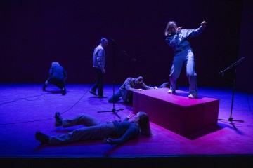 Viisi ihmistä esiintyy violetisti valaistulla lavalla, jossa on mikrofonit. Yksi henkilö seisoo ja elehtii tarmokkaasti korotetulla korokkeella, kun taas muut kyykistyvät, polvistuvat tai makaavat dramaattisesti lattialla luoden dynaamisen kohtauksen.