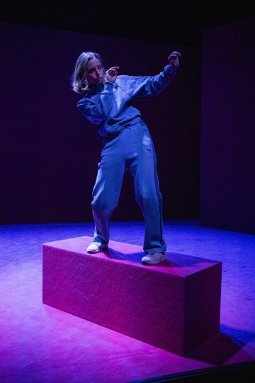Lyhyet vaaleat hiukset omaava, siniseen verkkareihin ja valkoisiin kenkiin pukeutunut henkilö seisoo vaaleanpunaisella suorakulmaisella alustalla ja poseeraa dynaamisesti dramaattisessa sinisessä ja vaaleanpunaisessa näyttämövalaistuksessa.