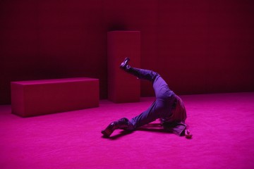 Tummiin vaatteisiin ja saappaisiin pukeutunut henkilö tekee pääpysty- tai tanssiasennon magentanvärisellä lavalla, jota ympäröivät samanväriset suorakulmaiset palikat. Hänen kasvonsa eivät näy.