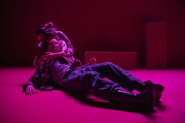 Kaksi ihmistä dramaattisessa valaistuksessa; toinen istuu lattialla taaksepäin lyyhistyneenä, ja toinen tukee häntä takaapäin syleillen. Kohtaus kylpee voimakkaassa punaisessa valossa, ja taustalla on vain vähän elementtejä.