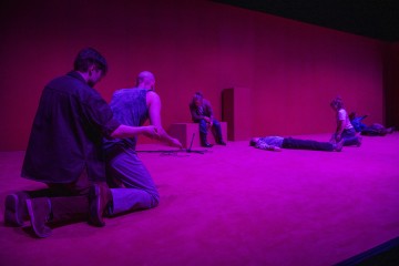 Seitsemän ihmistä on levittäytynyt violetissa valossa kylpevälle näyttämölle; jotkut polvistuvat, toiset istuvat tai makaavat lattialla, ja kaikki katsovat eri suuntiin synkkä tai mietiskelevä asenne.