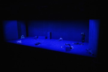 Hämärästi valaistulla, sinisessä valossa kylpevällä näyttämöllä näkyy useita ihmisiä, jotka makaavat tai istuvat lattialla eri asennoissa, ja heitä ympäröivät minimaaliset geometriset lavasteet. Yksi henkilö seisoo lähellä oikeaa reunaa ja elehtii.