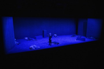 Syvänsinisellä valaistulla näyttämöllä on yksi henkilö, joka seisoo ja laulaa keskellä, ja hänen ympärillään on useita ihmisiä, jotka makaavat tai istuvat lattialla minimalistisessa, geometrisia muotoja sisältävässä lavastuksessa.