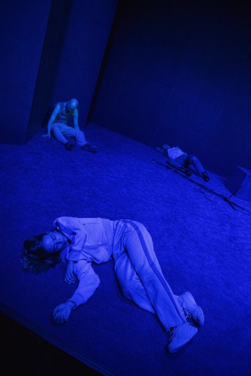 Kolme satunnaisesti pukeutunutta ihmistä makaa liikkumattomina lattialla ja seiniä vasten pimeässä huoneessa, jota valaisee sininen valo, joka luo dramaattisen ja synkän tunnelman.