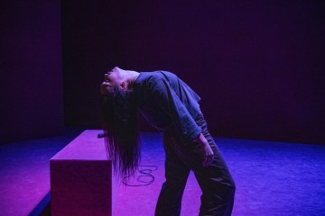 Pitkätukkainen henkilö nojaa dramaattisesti taaksepäin violetin ja vaaleanpunaisen näyttämövalaistuksen alla, lähellä suorakulmaista palikkaa ja lattialla olevaa mikrofonikaapelia.