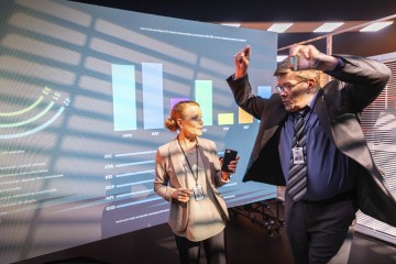 Nainen, jolla on kädessään puhelin, ja mies, joka nostaa kätensä ylös innostuneena, seisovat suuren digitaalisen näytön edessä, jossa näytetään värikkäitä pylväs- ja viivakaavioita modernissa toimistoympäristössä.