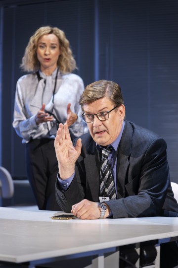 Pukumies nostaa kätensä pöydän ääressä istuen ja vakavana. Nainen seisoo hänen takanaan, elehtii käsillään ja puhuu, molemmat ammattimaisessa toimistoympäristössä.