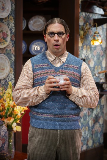Mies, jolla on silmälasit, sininen neuleliivi ja raidallinen paita, pitelee teekuppia molemmilla käsillään. Hän seisoo vanhassa, kukkakoristeisessa huoneessa, jossa lautaset ovat hyllyillä ja kukkakimppu hänen vieressään.