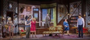 Värikäs lavastus vintage-olohuoneesta, jossa on kuusi aikakauden pukuihin pukeutunutta näyttelijää, joista osa elehtii tai puhuu vilkkaasti. Huoneessa on kukkatapetti, vintage-huonekaluja sekä erilaisia koriste-esineitä ja maalauksia.