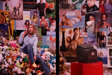 Yllättynyt henkilö istuu pehmolelujen keskellä huoneessa, joka on täynnä elokuvien ja julkkisten julisteita, joita jakaa musta viiva. Etualalla on pieni televisio punaisella pöydällä ja valkoinen lamppu.