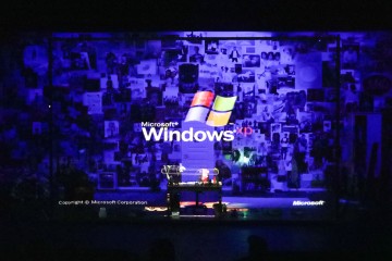 Hämärästi valaistussa huoneessa on kollaasipeitteinen seinä ja Windows XP:n käynnistysnäyttö, joka heijastetaan työpöydän yläpuolelle, jossa istuu henkilö tietokoneen ääressä, mikä luo nostalgisen digitaalisen tunnelman.