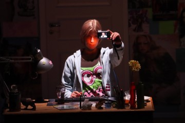 Vaaleahiuksinen henkilö istuu hämärässä huoneessa sekavan työpöydän ääressä ja pitää puhelinta ylhäällä ottaakseen selfietä. Hänellä on yllään vaaleansininen huppari ja värikäs paita, jossa on sarjakuvamalli. Julisteet ja valokuvat peittävät seinää heidän takanaan.