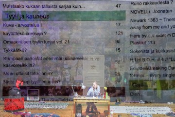 Henkilö istuu ja työskentelee luovassa tilassa sotkuisen pöydän ääressä, jonka päällä on puoliksi läpinäkyvä lista, jossa on suomenkielistä tekstiä, numeroita ja aikakauslehtityylisiä otsikoita. Taustalla näkyy julisteita ja erilaisia taidemateriaaleja.