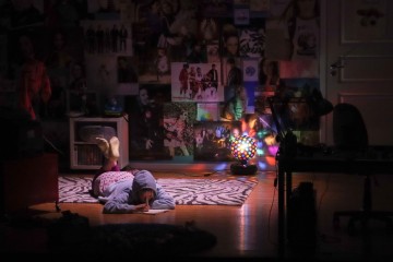 Henkilö makaa vatsallaan piirtäen lattialla hämärästi valaistussa, julisteilla päällystetyssä huoneessa. Värikäs diskopallolamppu heittää värikkäitä valoja, jotka lisäävät eloisaa ja kodikasta tunnelmaa.