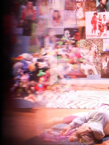 Sumeassa kuvassa näkyy etualalla lattialla makaava henkilö, jonka takana seinää peittävät värikkäät julisteet ja pehmolelut. Kohtaus vaikuttaa kaoottiselta ja eloisalta, ja siinä on kirkas ja pehmeä valaistus.